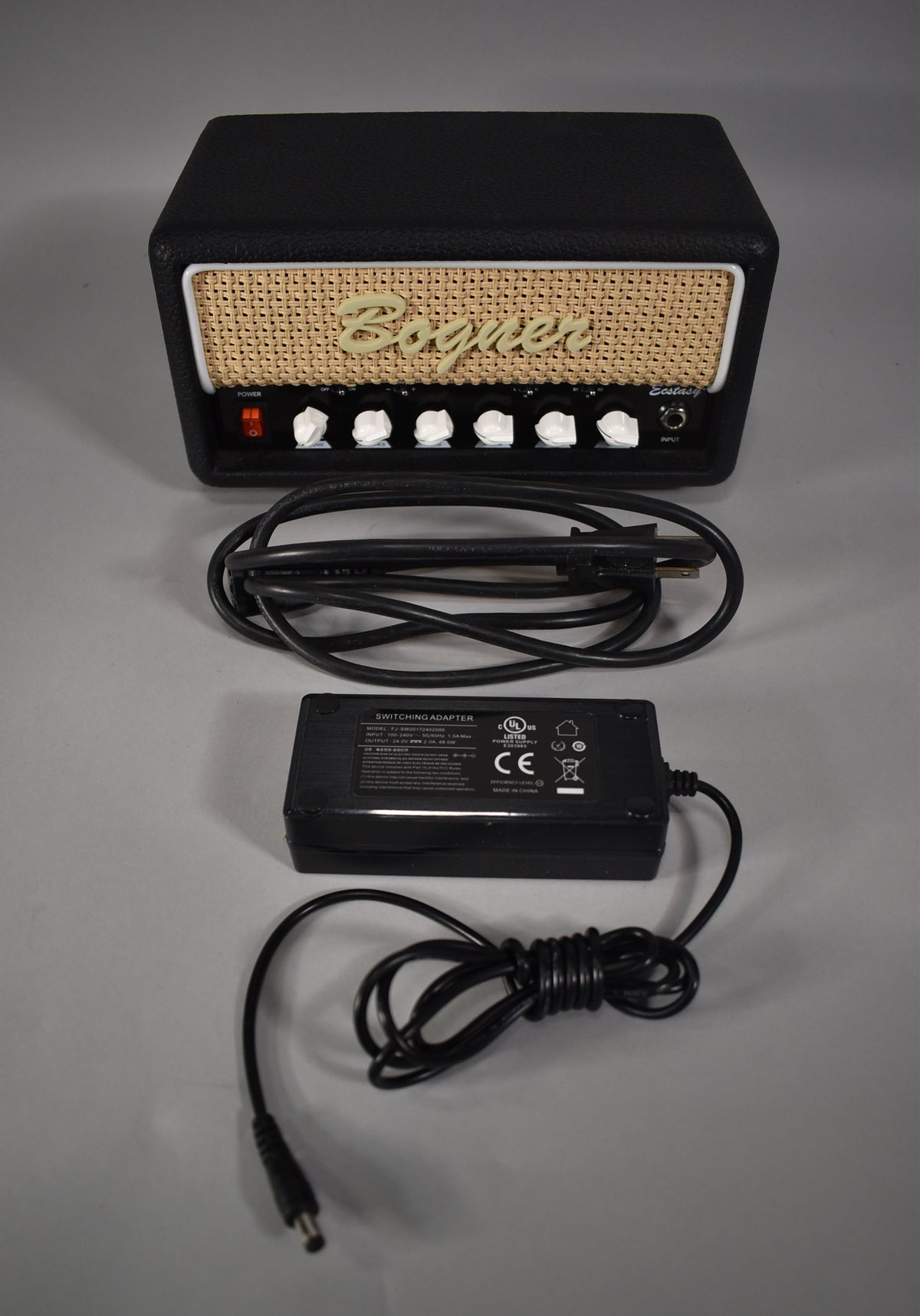 Black amp head with a tan woven grille, six white knobs, a power switch, and an input jack, shown with its power supply.