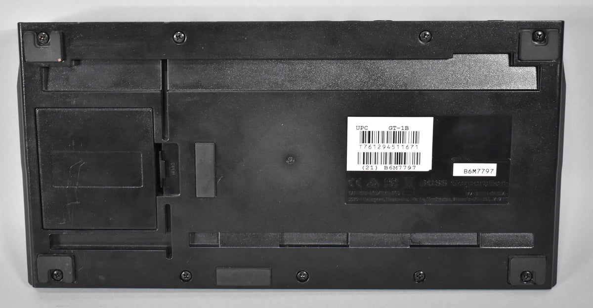 The black plastic pedal is seen from the bottom, showing its battery compartment, four rubber feet, and a barcode sticker.