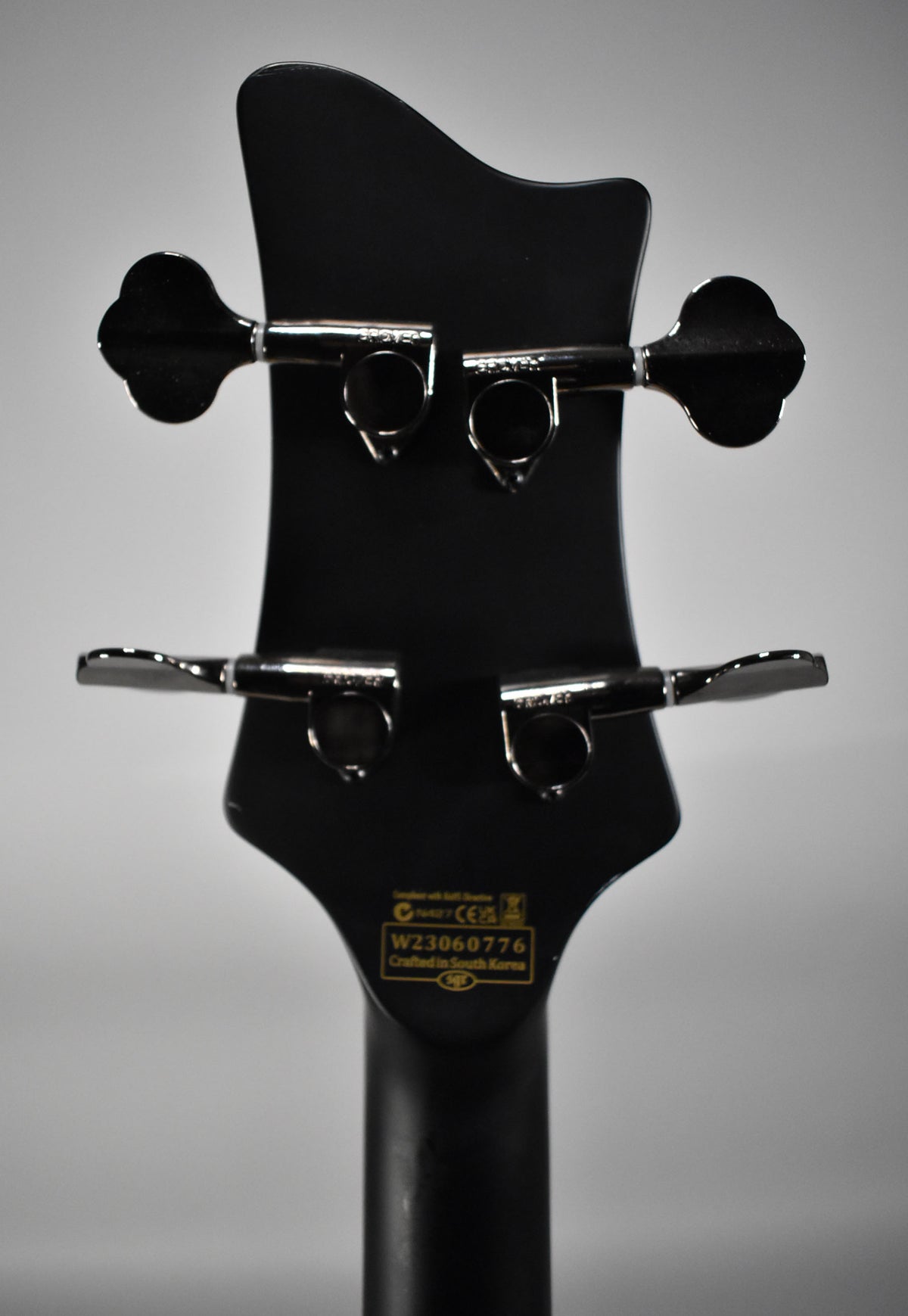 The back of the bass headstock, matte black with four chrome tuning machines, and a yellow serial number sticker.