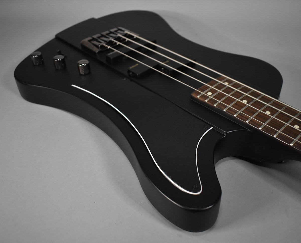 Angled close-up of the matte black bass body, showing three chrome knobs, two pickups, and a white pinstripe along the lower edge.