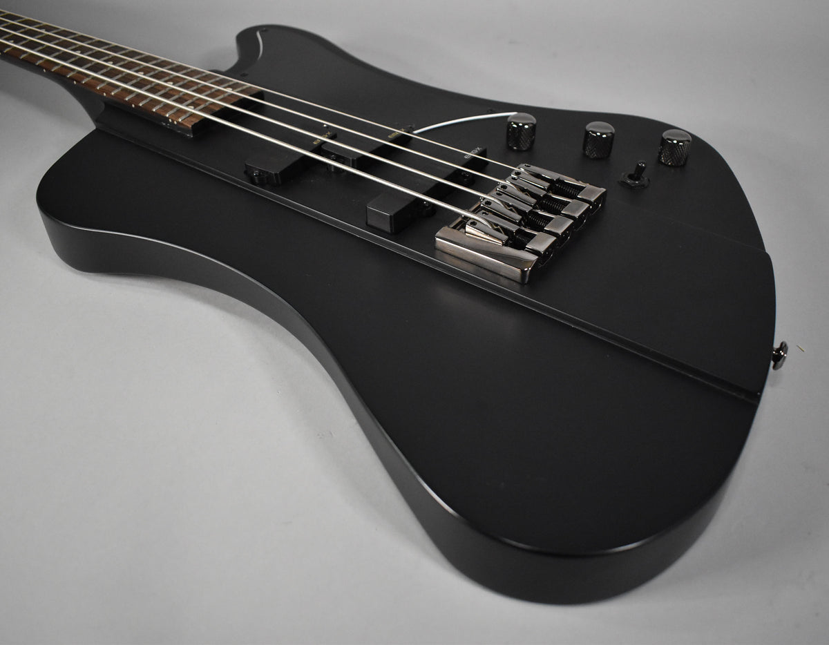 Close-up of the satin black bass body, showing the chrome bridge, two black pickups, three knurled knobs, and a toggle switch.