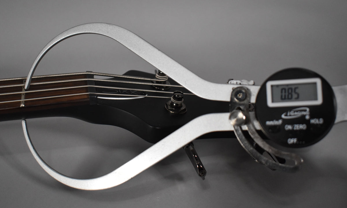 Close-up of the bass's black satin headstock and dark fretboard, with a digital caliper measuring string height at 0.85mm.