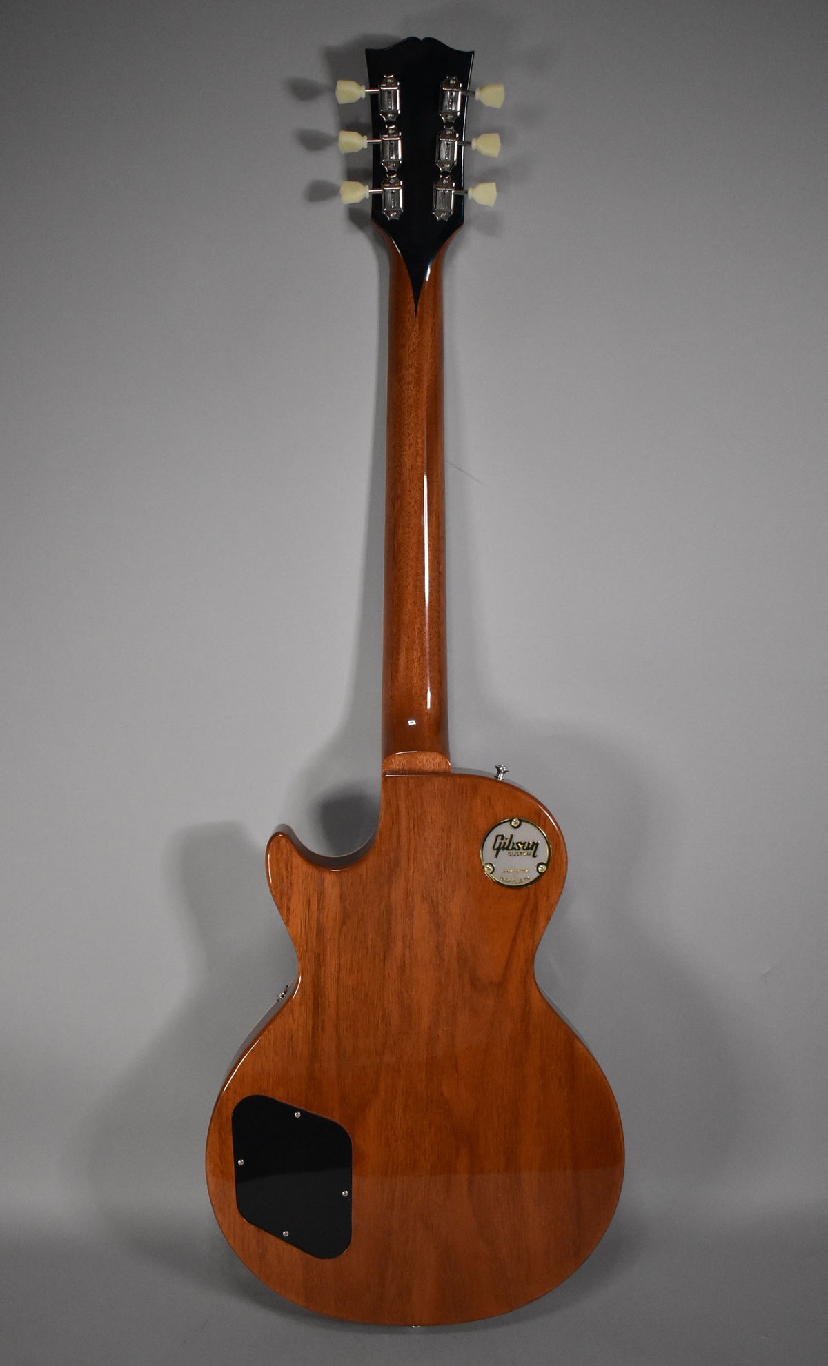 Full back view of a glossy, light brown Les Paul with a black headstock and cream tuning pegs.