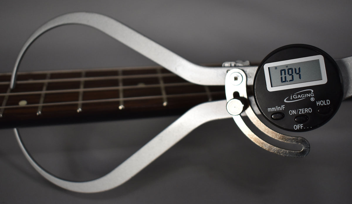 A digital caliper measures the bass's dark fretboard, showing "0.94" on its screen.