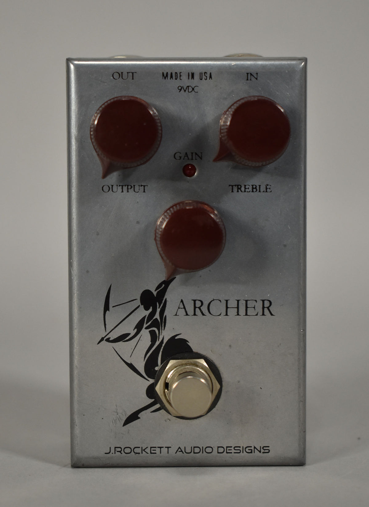 A brushed silver pedal, viewed from above, with three dark red pointer knobs, a red LED, and a black archer graphic.