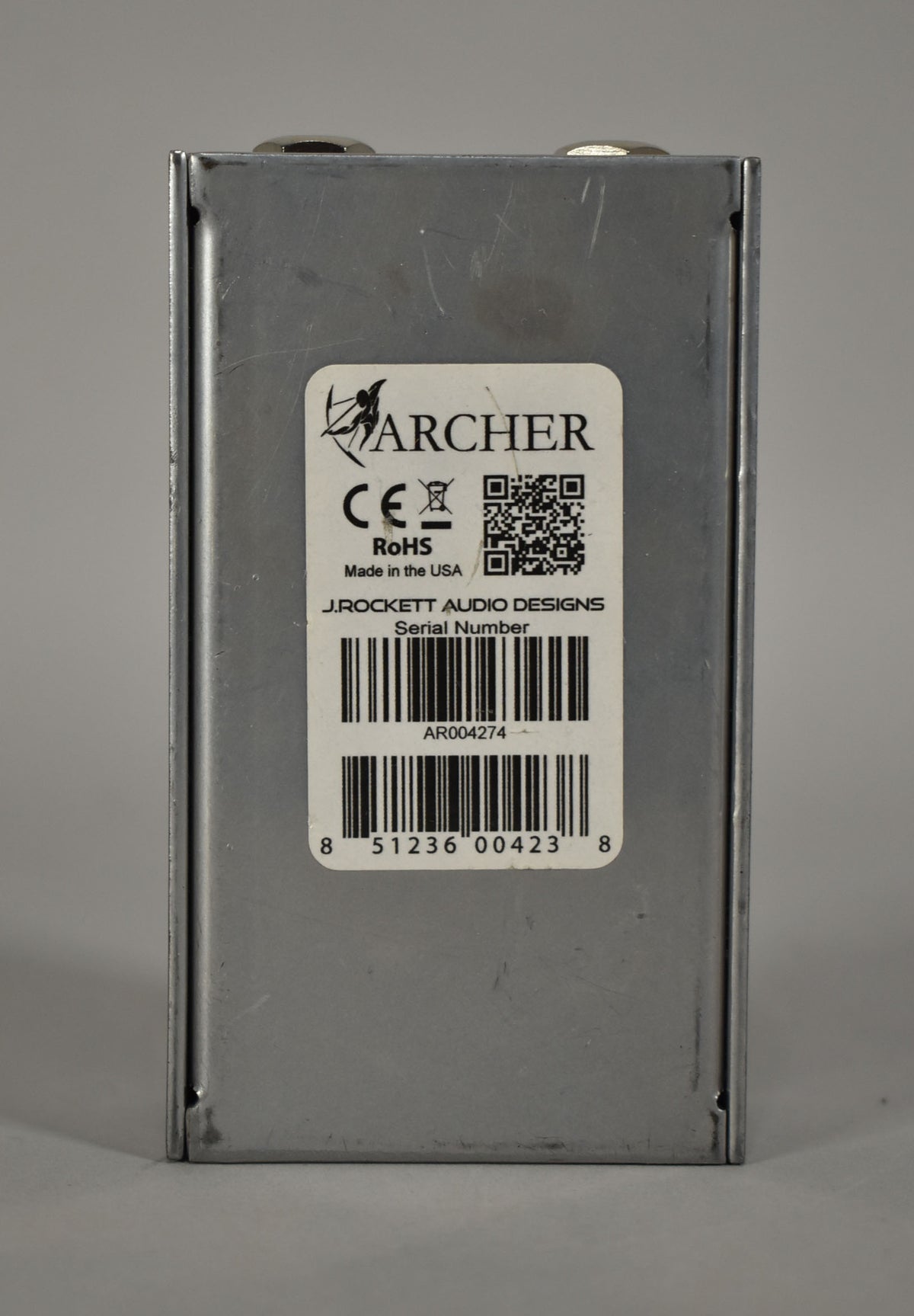 The pedal's silver metal casing is viewed from the back, showing a white label with a barcode and serial number.