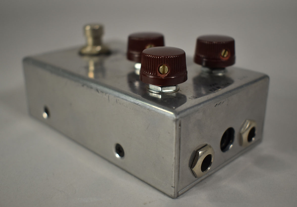 The brushed aluminum pedal is angled right, showing three dark brown ridged knobs, a footswitch, and two input jacks.