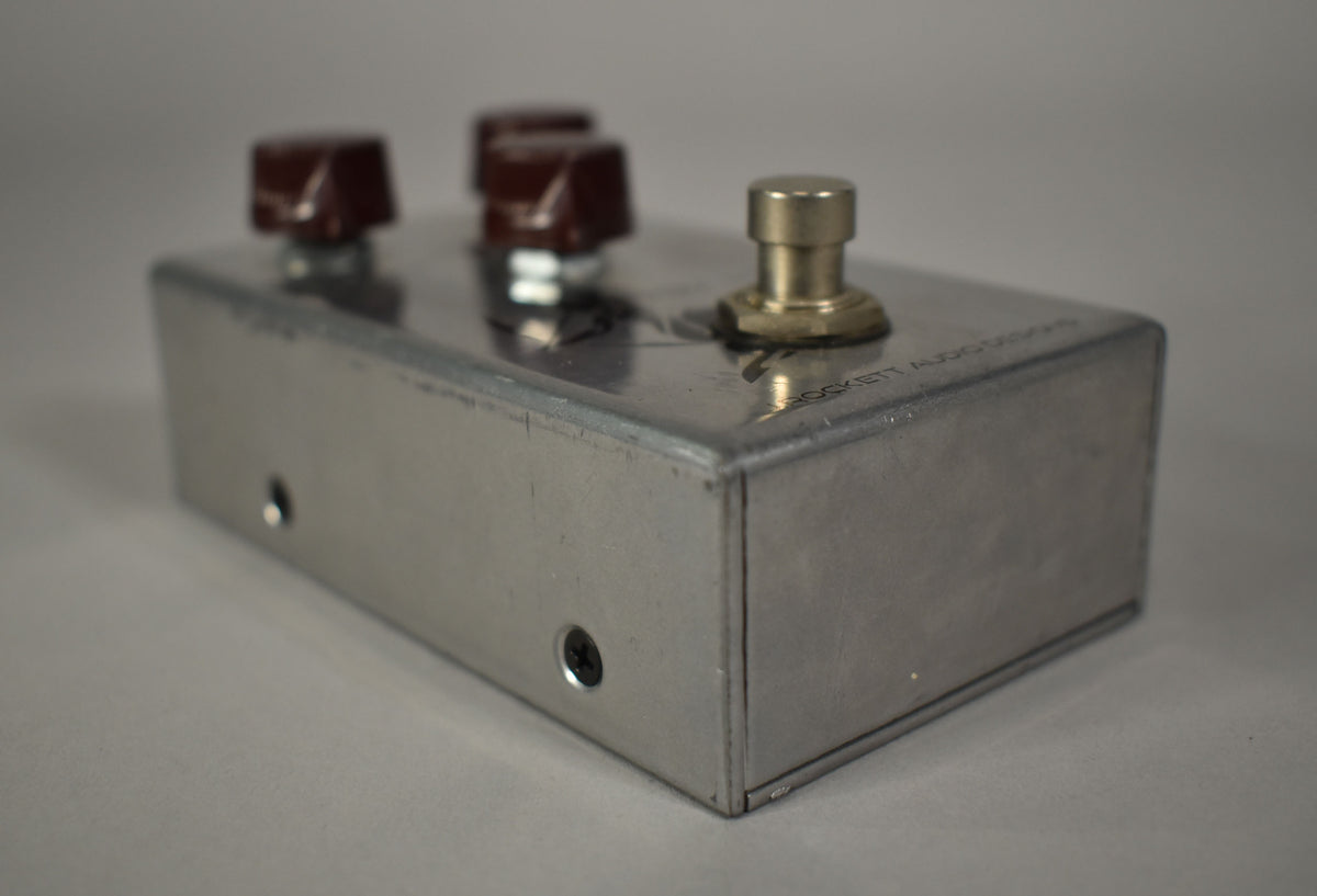 The brushed aluminum pedal is angled from the left, showing three dark brown knobs, a brass footswitch, and minor scuffs.