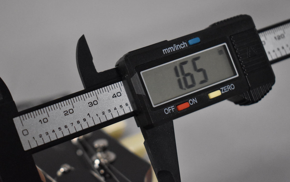 A digital caliper measures the nut width of the guitar at 1.65 inches, with the headstock and tuning pegs visible below.