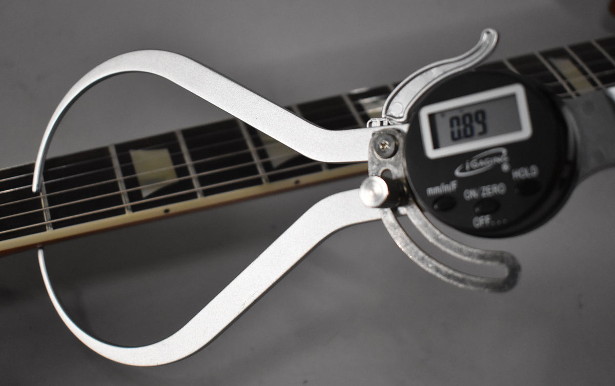 A digital caliper measures the guitar's fretboard, showing "0.89" on its display, with trapezoid inlays visible on the neck.