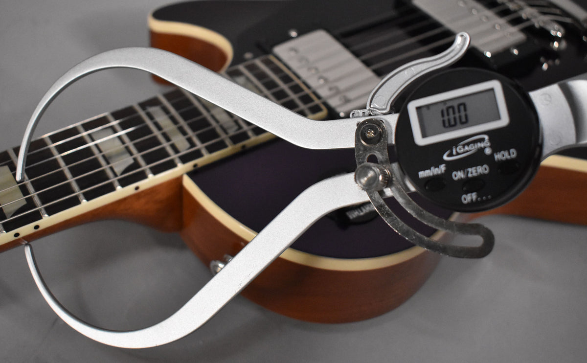 A digital caliper measures the neck thickness of the black Les Paul, showing "1.00" on its display.
