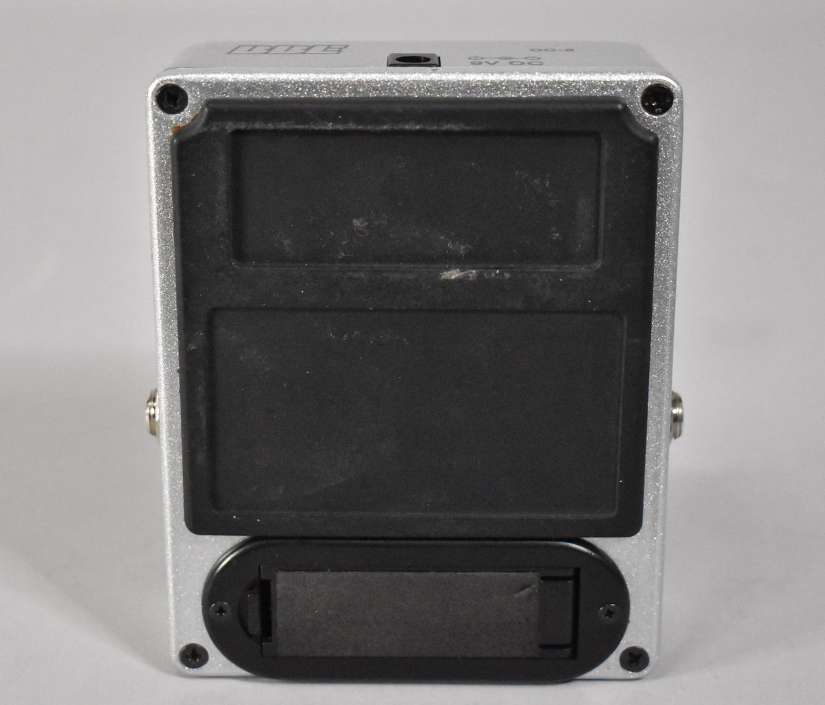 The silver pedal is viewed from the back, showing its black baseplate with two large rubber pads and a battery compartment.