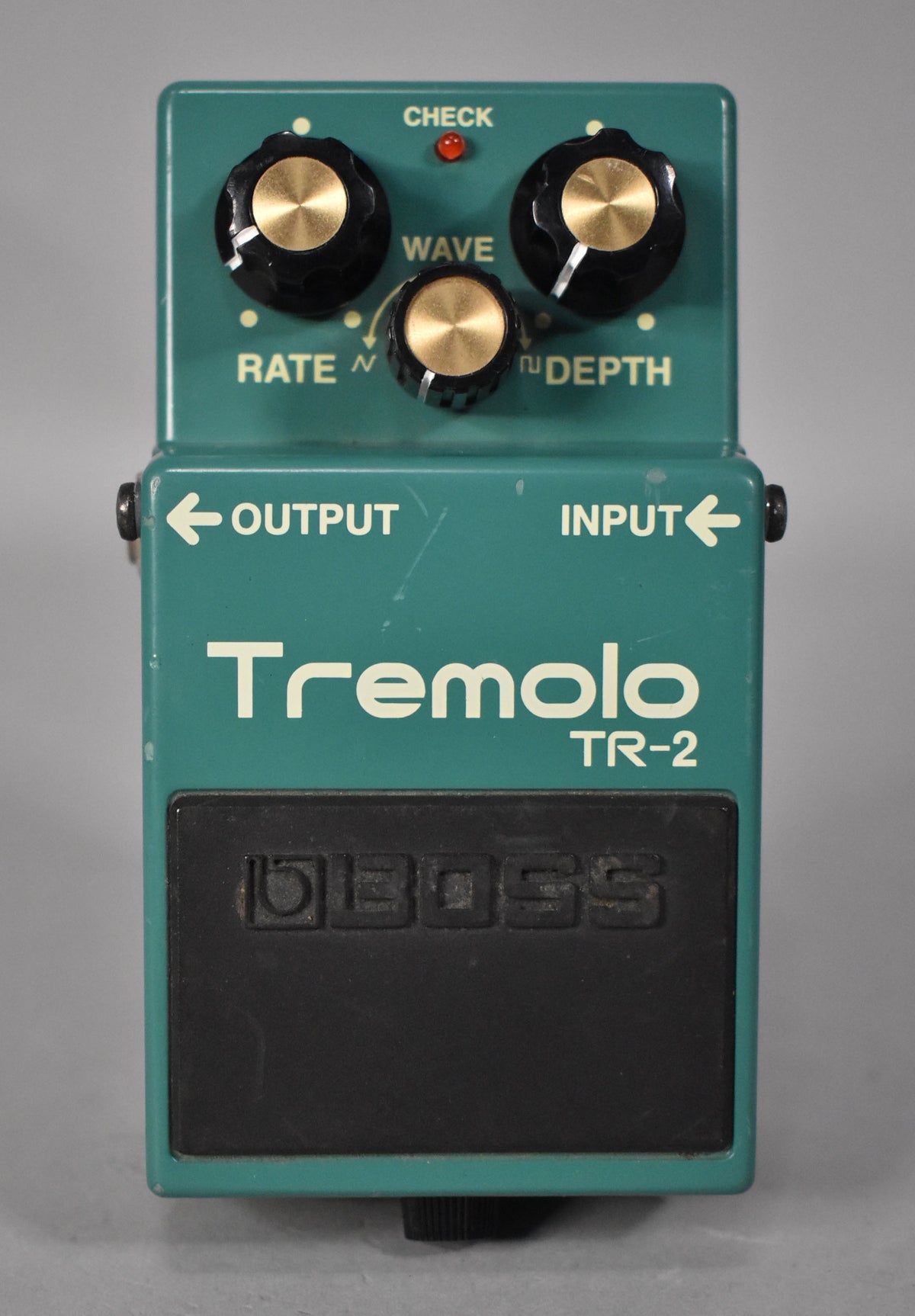 A teal green guitar pedal, viewed from above, with three black and gold knobs, a red LED, and a black footpad.