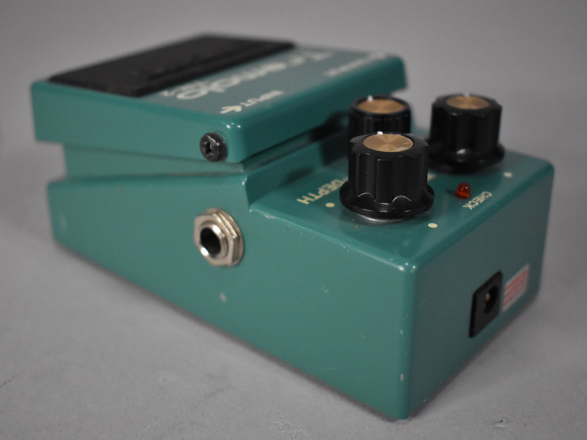 A teal-green pedal, angled right, showing the input jack, three black knobs with gold tops, and a red indicator light.