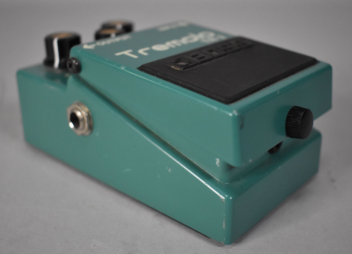 A teal pedal, viewed from the left, shows a silver input jack, two black knobs, and a black footswitch with visible scuffs.