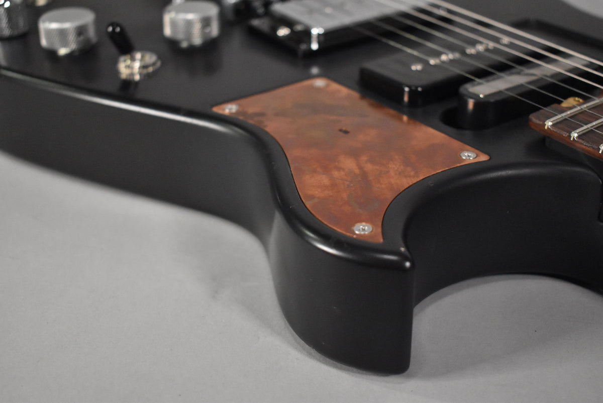Close-up of the matte black guitar body, showing a distressed copper pickguard, two knurled chrome knobs, and a toggle switch.