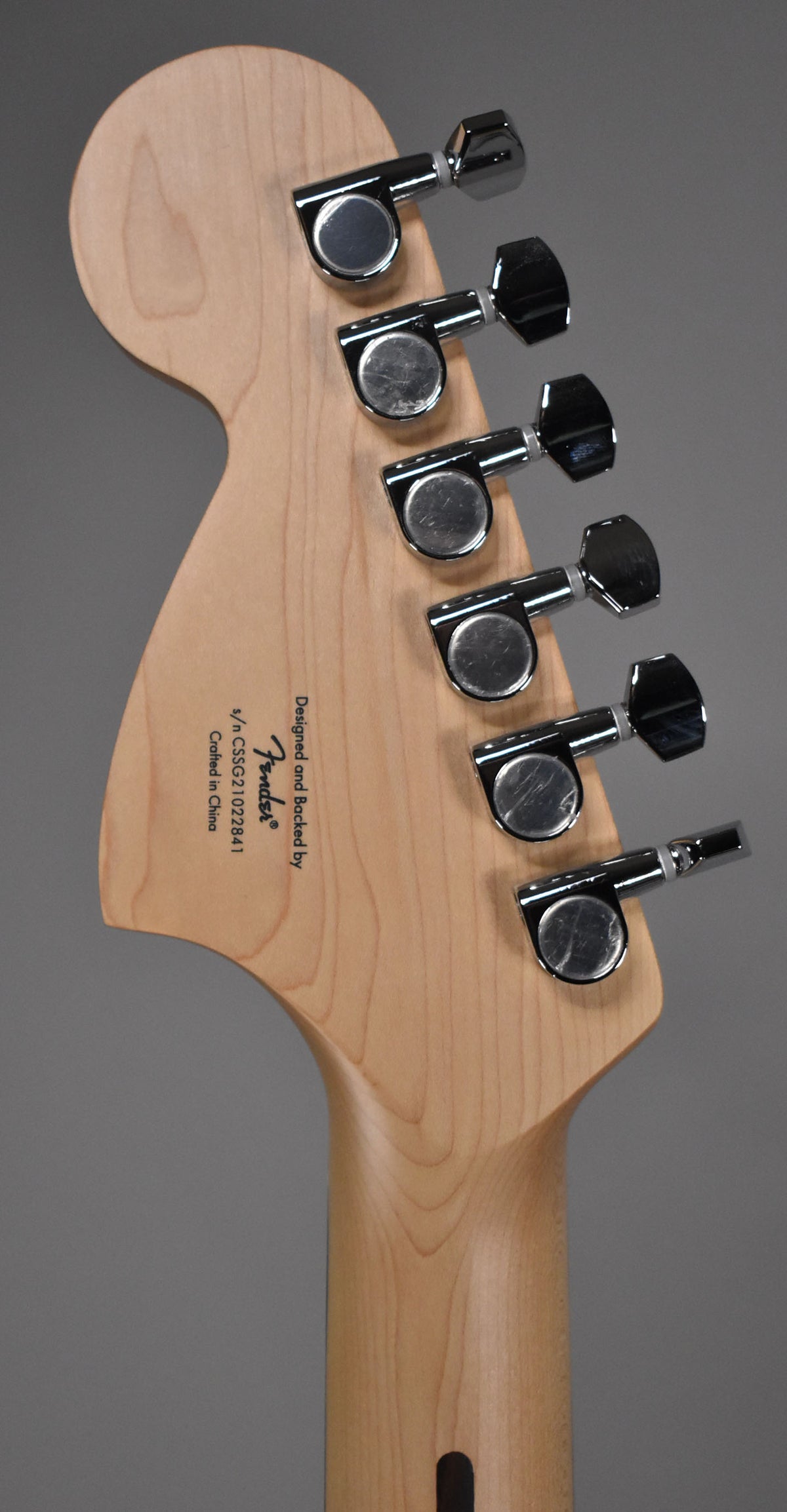 The back of a light wood headstock and neck, angled to show six chrome tuning machines with rectangular buttons.