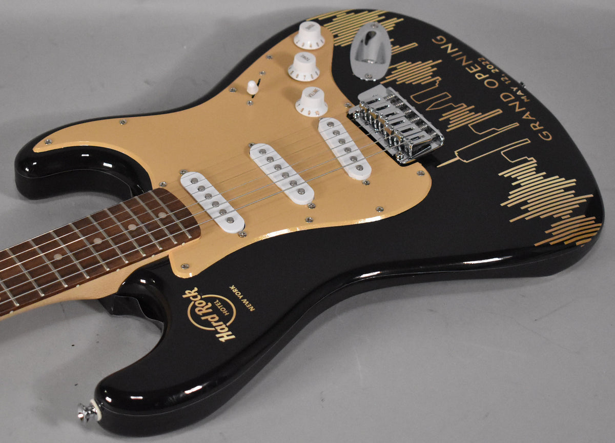 Black Stratocaster with a gold pickguard, white pickups and knobs, and gold Hard Rock Cafe NYC graphics on the body.