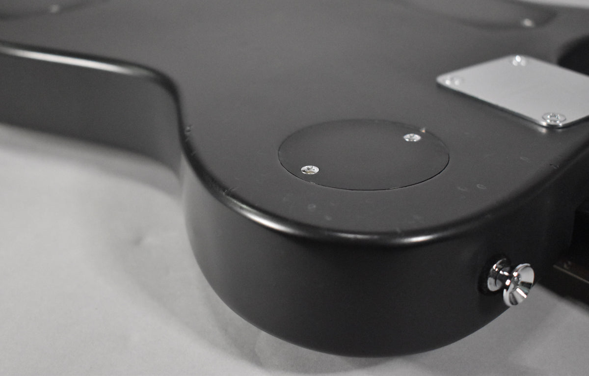 Close-up of the guitar's matte black body, showing a circular control plate, a rectangular plate, and a chrome strap button.