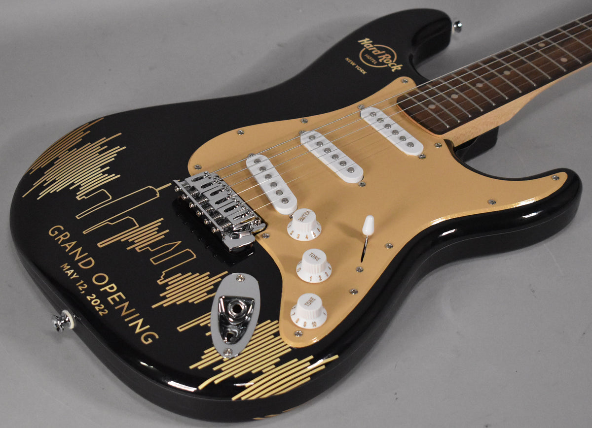 Black Stratocaster with a gold pickguard, white pickups, and a gold skyline graphic with "Grand Opening May 12, 2022" text.