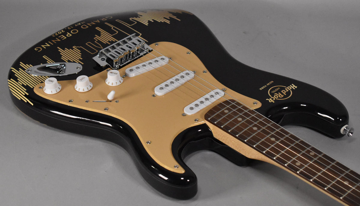 Black Stratocaster with a gold pickguard, three white single-coil pickups, and gold Hard Rock Cafe NYC graphics on the body.