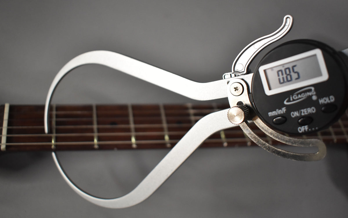 A digital caliper measures the guitar's dark fretboard, showing "0.85" on its screen, with strings and frets visible.