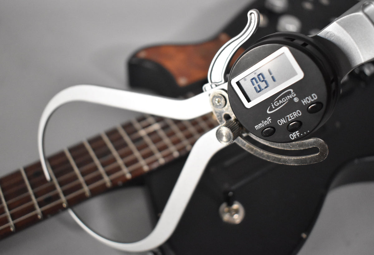 A digital caliper measures the fret height on the dark fretboard of a black electric guitar, displaying "0.91".