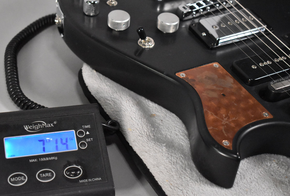 Close-up of the guitar's lower body, showing a relic black finish, copper control plate, and a scale displaying 7 lbs 14 oz.
