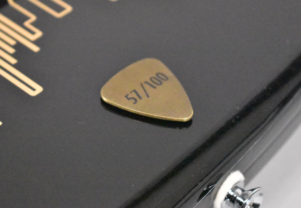 Close-up of the black guitar body, showing a gold pick engraved "57/100" and a chrome strap button with a white felt washer.