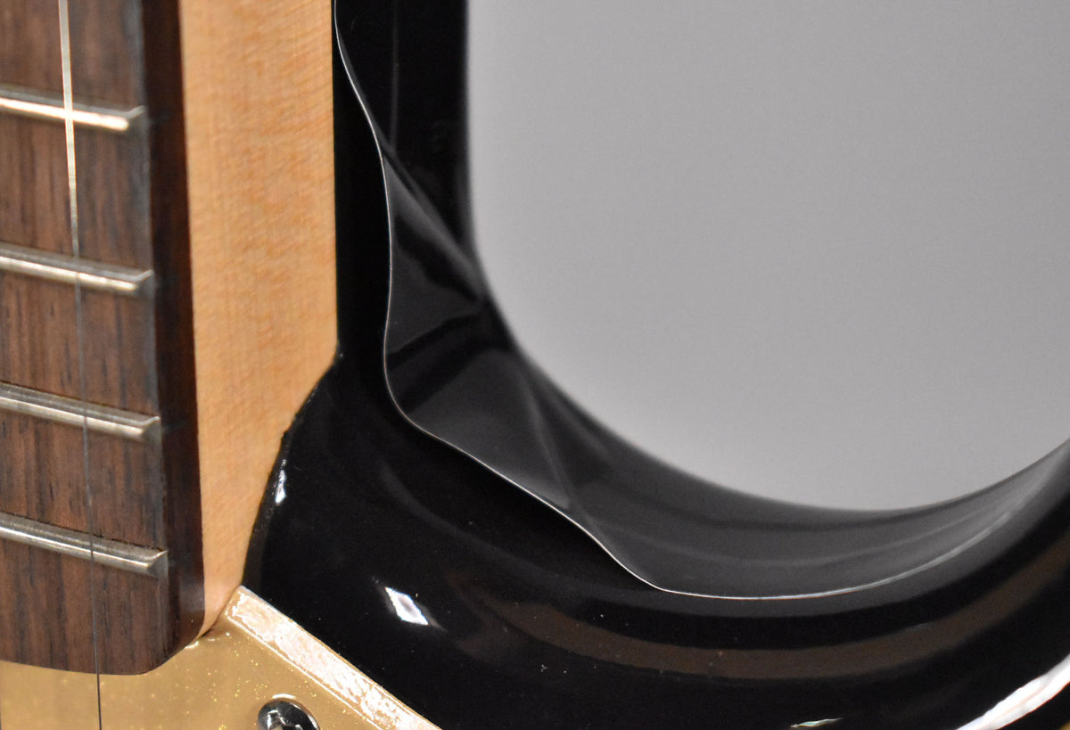 Close-up of the guitar's black body and neck joint, showing a partially peeled protective film and a small chip in the finish.