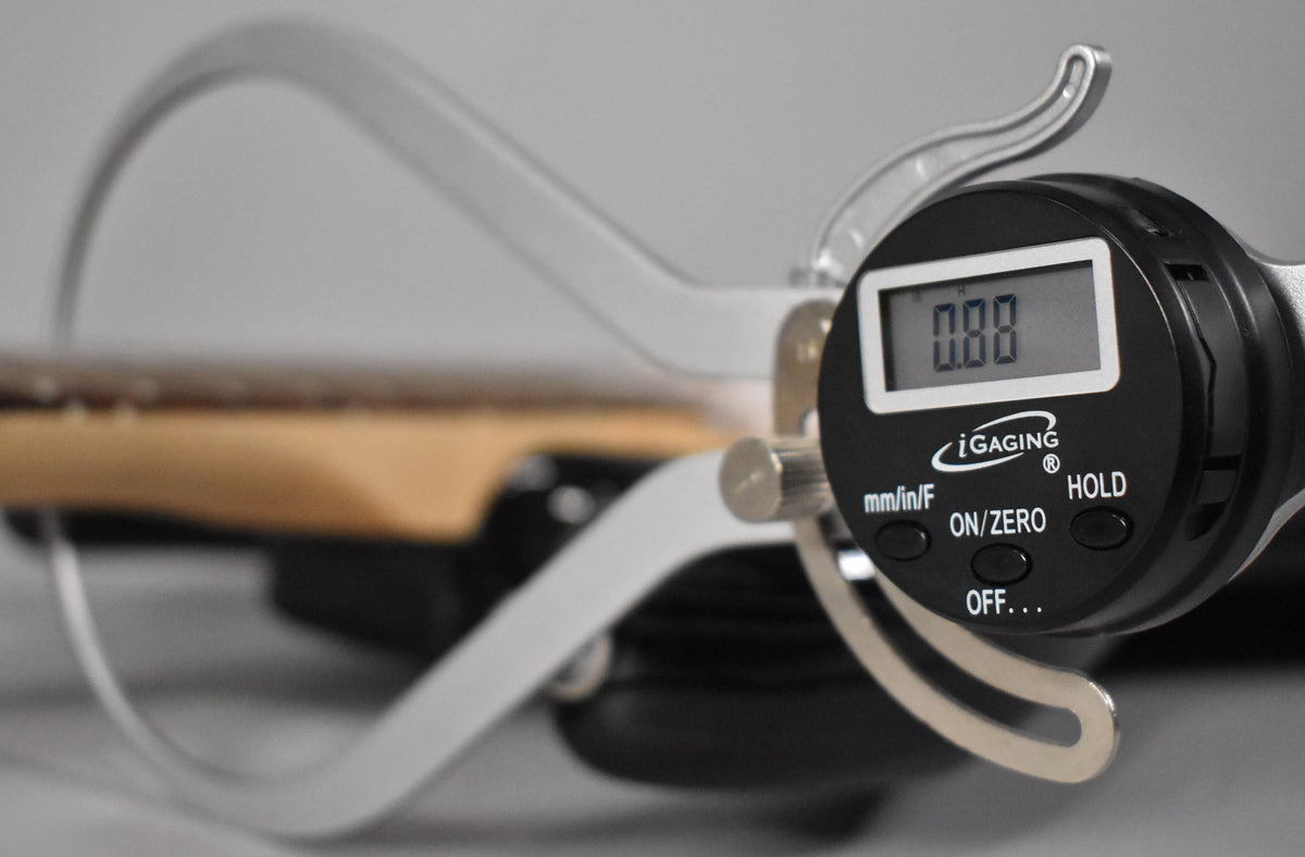 A digital caliper measures the guitar's light-colored neck, showing "0.00" on its display.