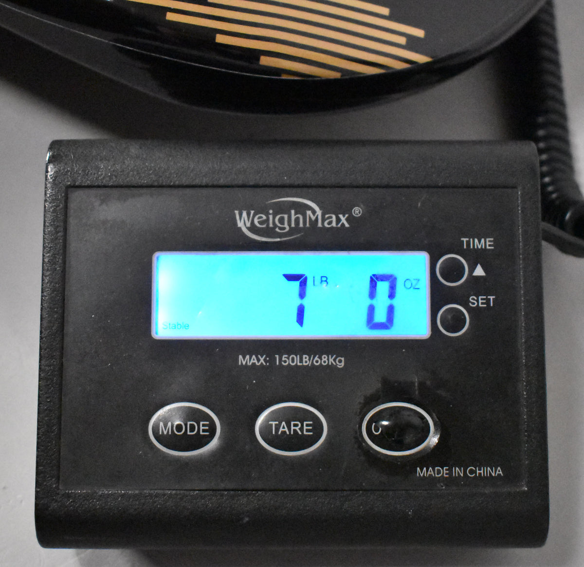 A close-up of a digital scale displaying "7 LB 0 OZ" in blue, with a guitar's black body partially visible above.