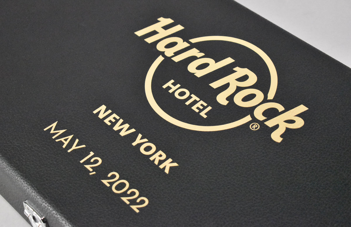 Close-up of a black textured guitar case with gold Hard Rock Hotel New York and May 12, 2022 lettering, and a silver latch.