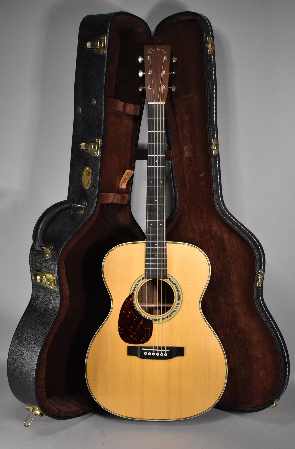 Full-length view of a natural finish acoustic guitar with a tortoiseshell pickguard, standing upright in its open, dark brown hard case.