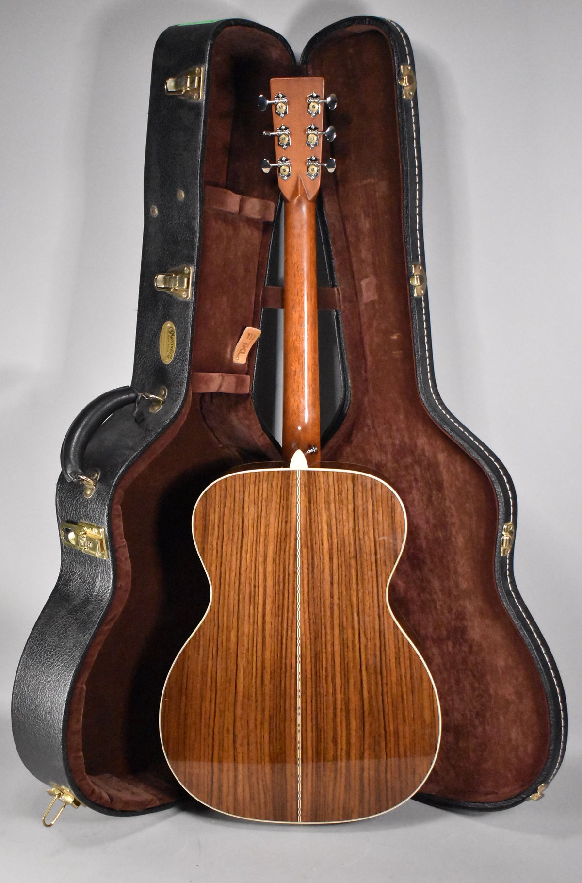 The guitar's back and neck are visible, with a glossy natural finish and prominent wood grain, resting in an open hard case.