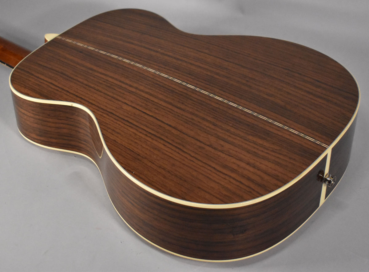 The back of the dark brown acoustic guitar, with a light binding, is angled to show the wood grain and a visible endpin jack.