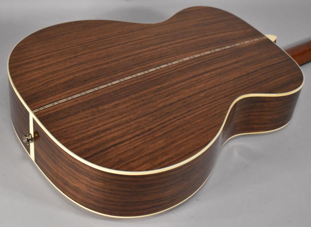 The guitar's dark brown back and sides, with a prominent grain and light binding, are angled to show the endpin jack.