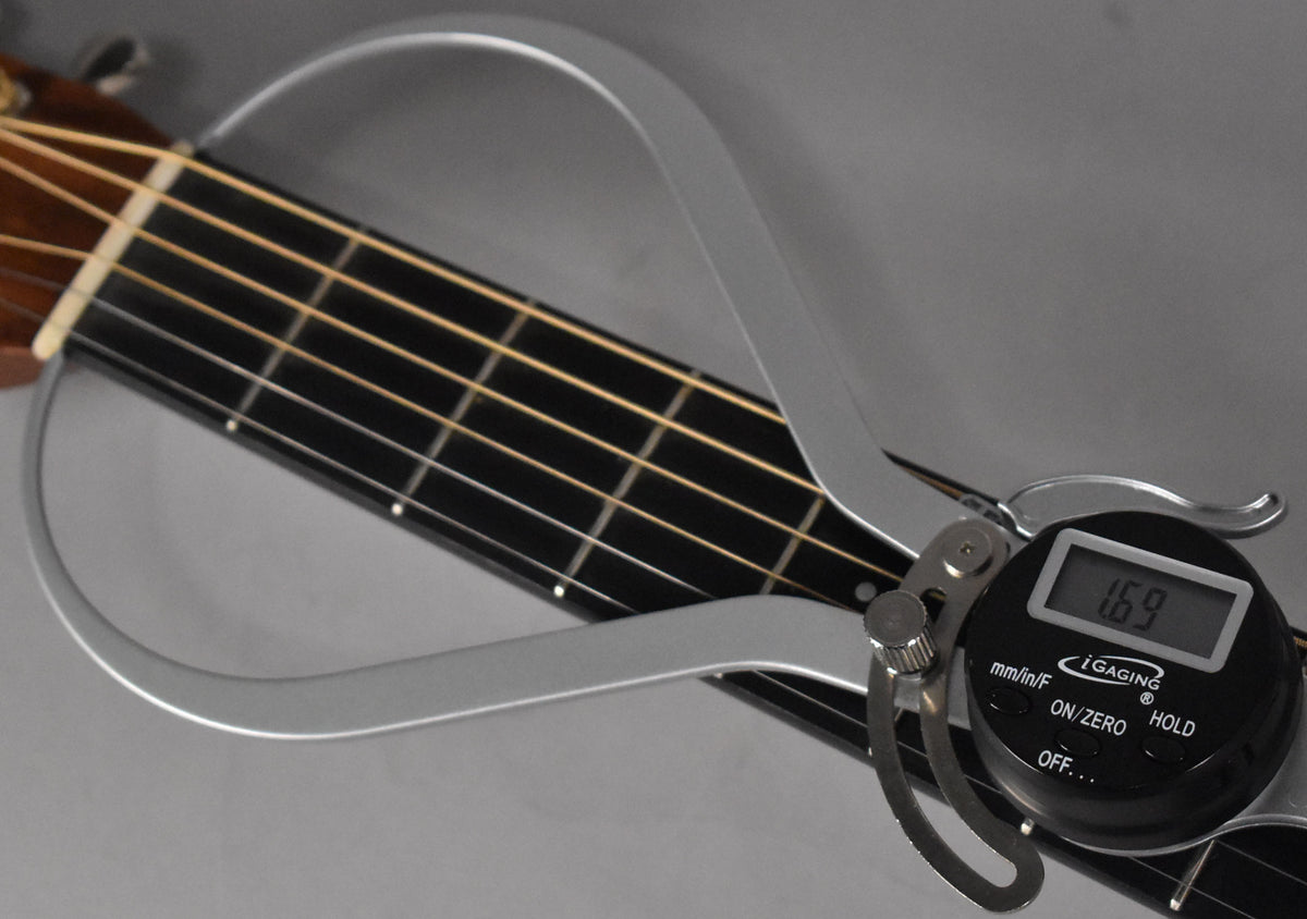 A digital caliper measures the nut width of the natural finish acoustic guitar, showing 1.69 inches on its display.