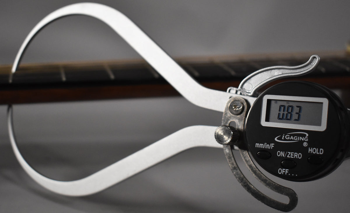 A digital caliper measures the guitar's fretboard thickness, displaying "0.83" on its screen, with the dark brown fretboard visible.