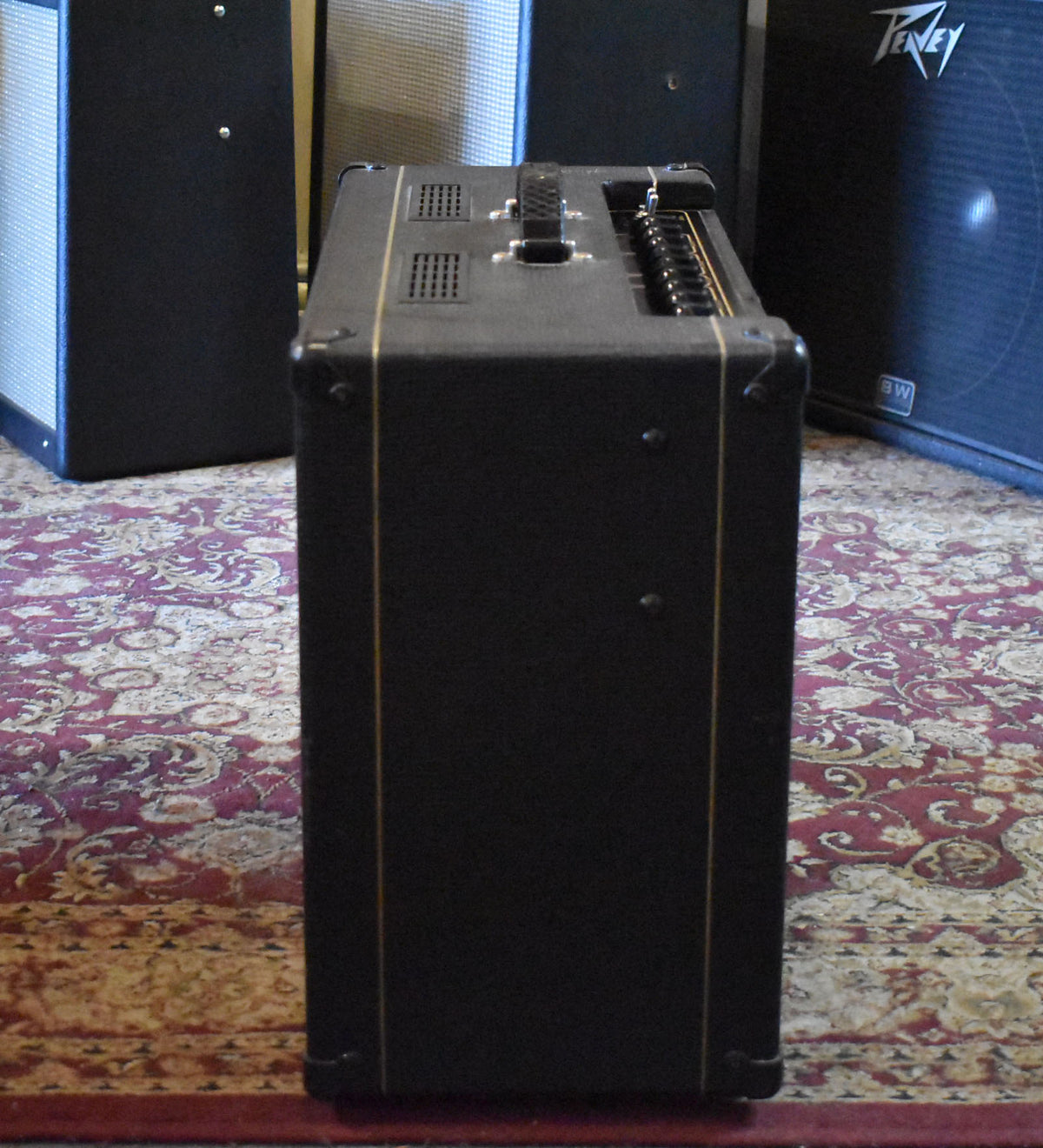 The black combo amp is seen from its right side, showing the top control panel with many black knobs and a central carrying handle.