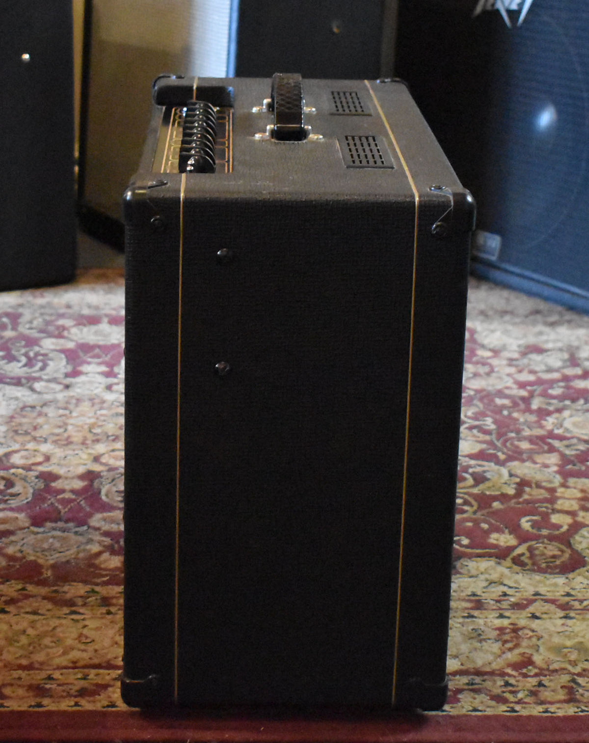 The black textured amp is viewed from the right side, showing a top handle, a row of black knobs, and two rectangular vents.