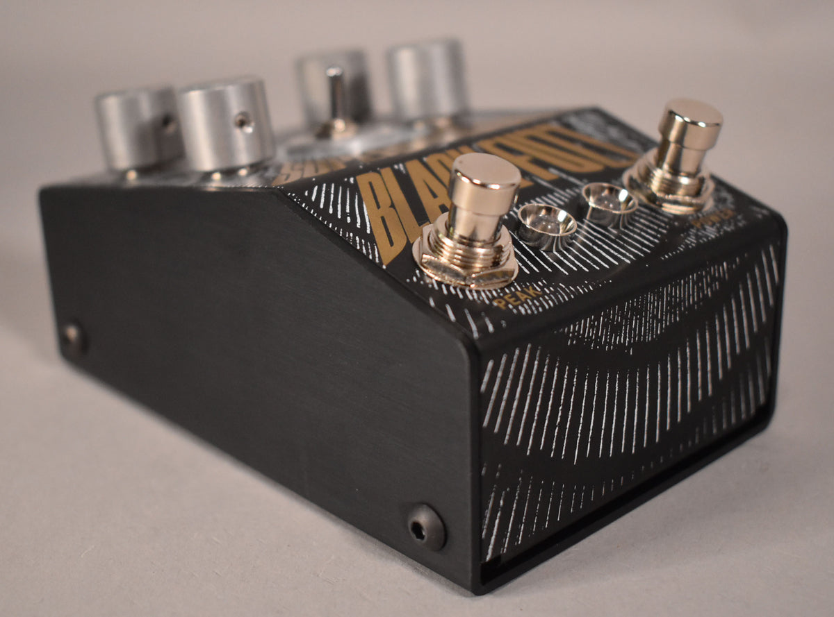 A black fuzz pedal with gold and silver graphics, viewed from the front right, showing two gold footswitches and four silver knobs.
