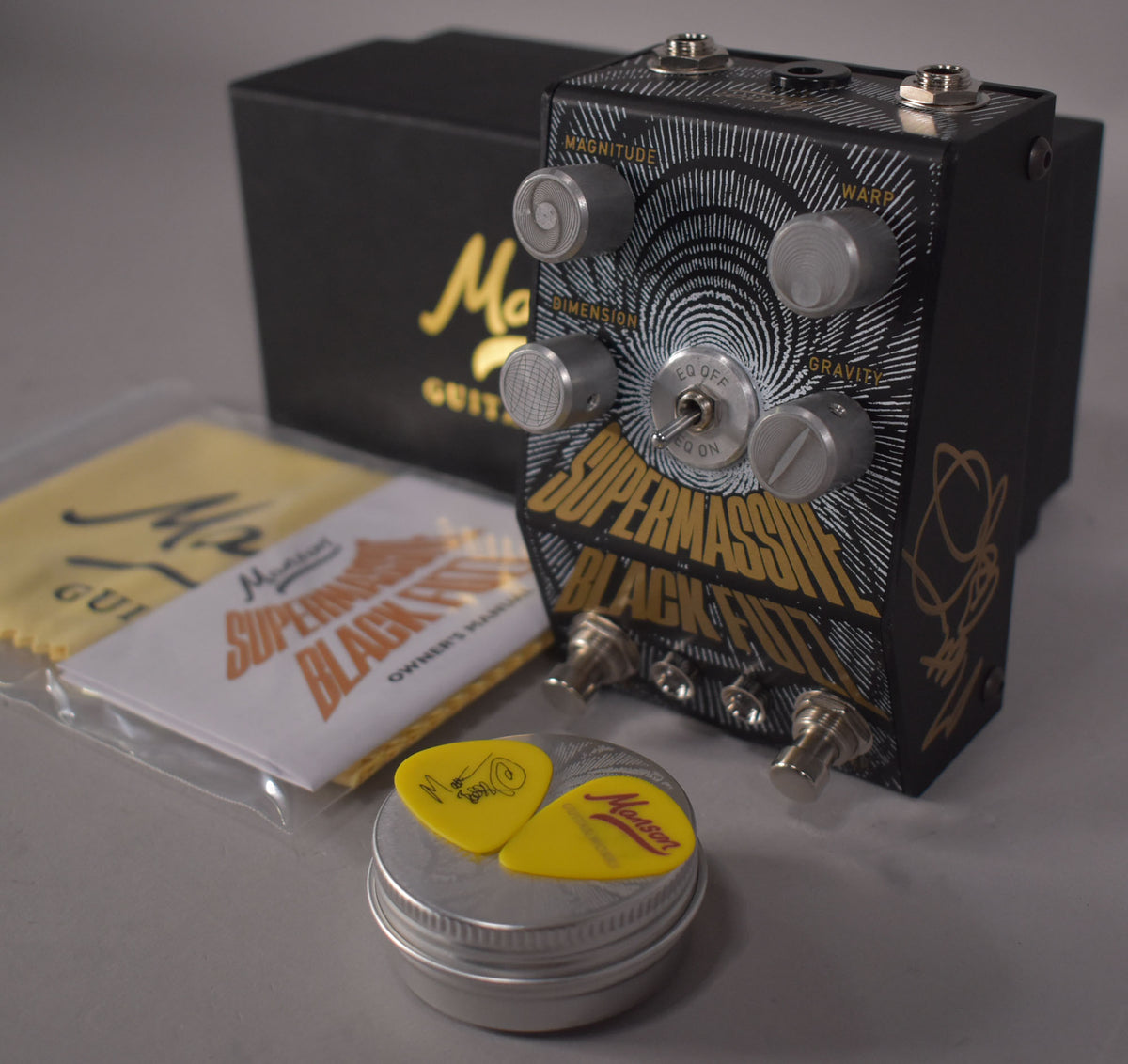 A black fuzz pedal with gold lettering and white concentric circles, four silver knobs, and a toggle switch, shown with its box and accessories.