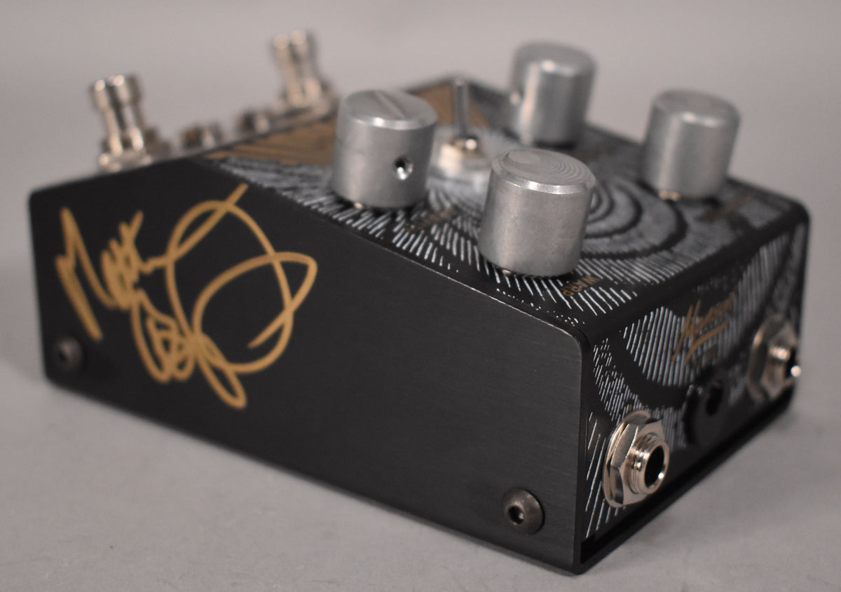 The black pedal is angled right, showing a gold signature on the left side and four silver knobs on the top, with two input jacks on the right.