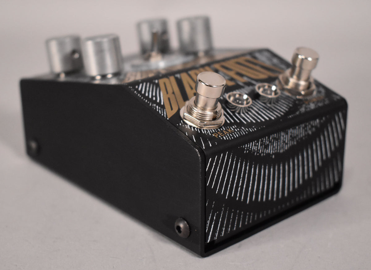 A black guitar pedal with gold and white graphics, viewed from the right, showing three silver knobs and two footswitches.