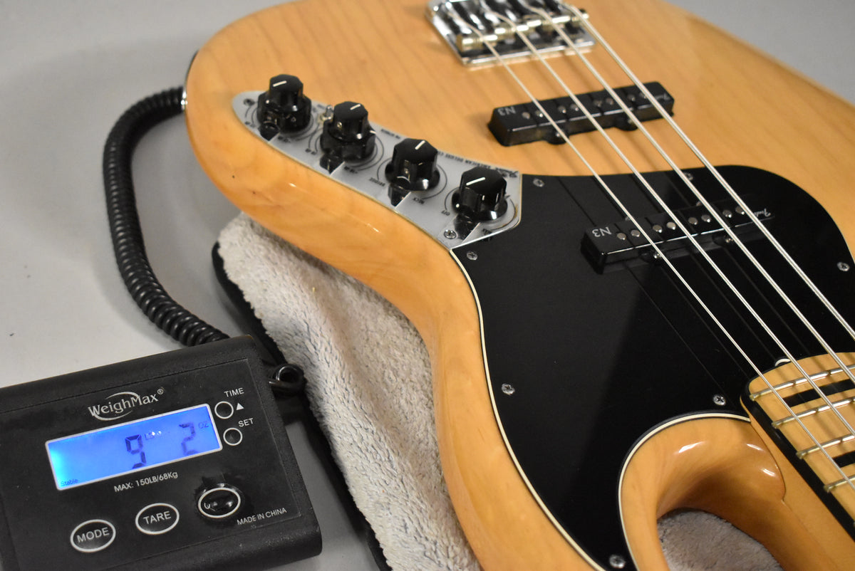The natural finish bass, with a black pickguard and four black control knobs, rests on a towel next to a scale displaying "9.2".