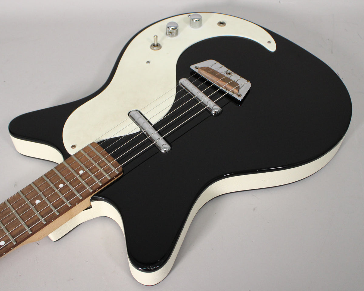 Close-up of a black Danelectro DC-59 guitar with a white pickguard, showing the body, bridge, pickups, controls, and part of the fretboard.