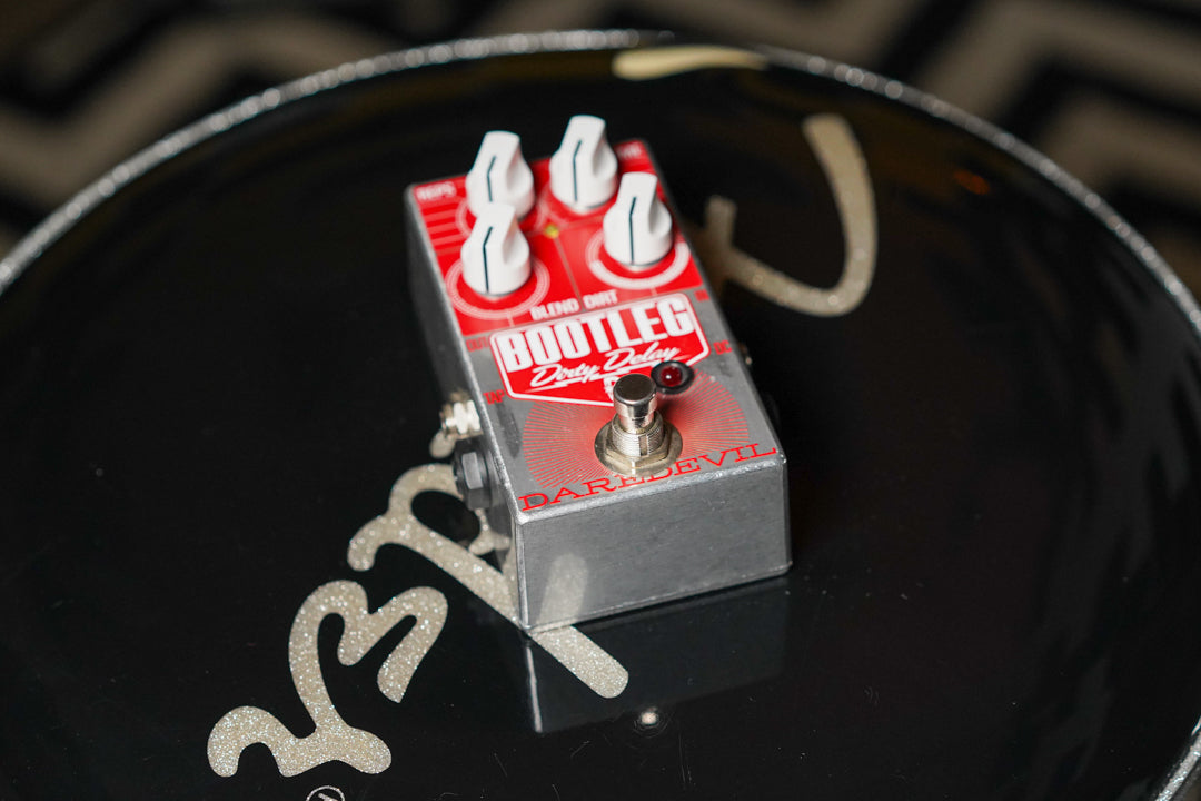 The silver-bodied pedal, with a red and white faceplate, is angled from above, showing four white knobs and a silver footswitch.