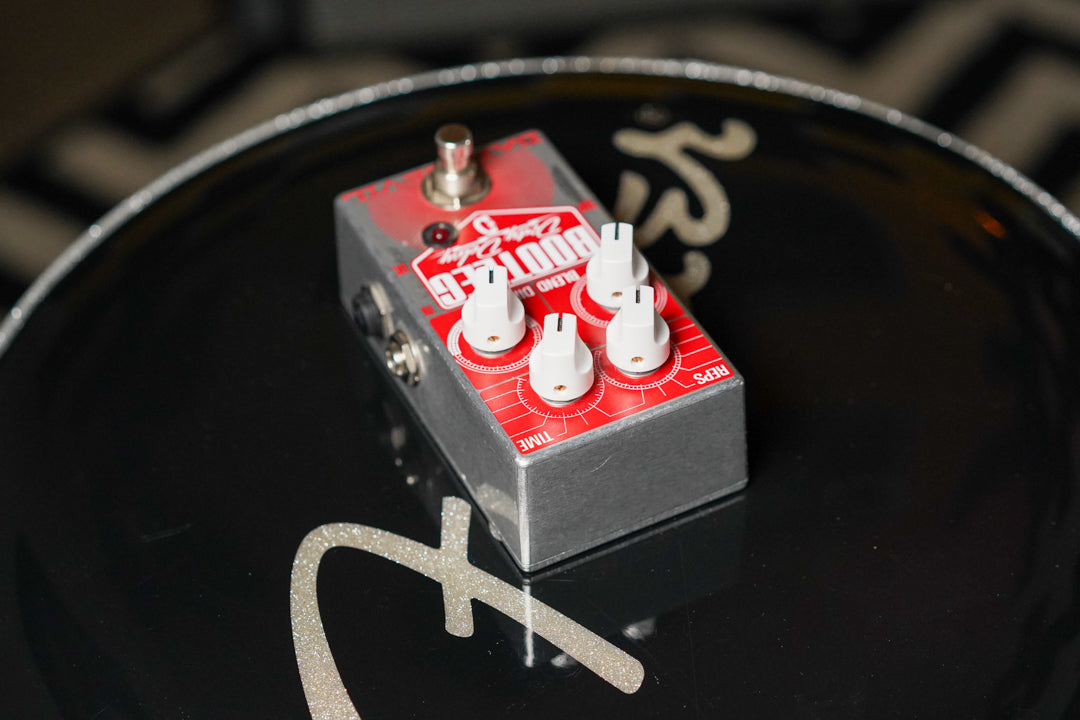 The silver pedal with a red faceplate and four white knobs is angled, showing a side input jack and top footswitch.