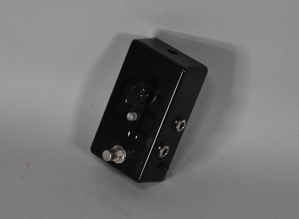 A glossy black rectangular pedal, viewed from the top right, with a large black knob, a small silver knob, and a silver footswitch.